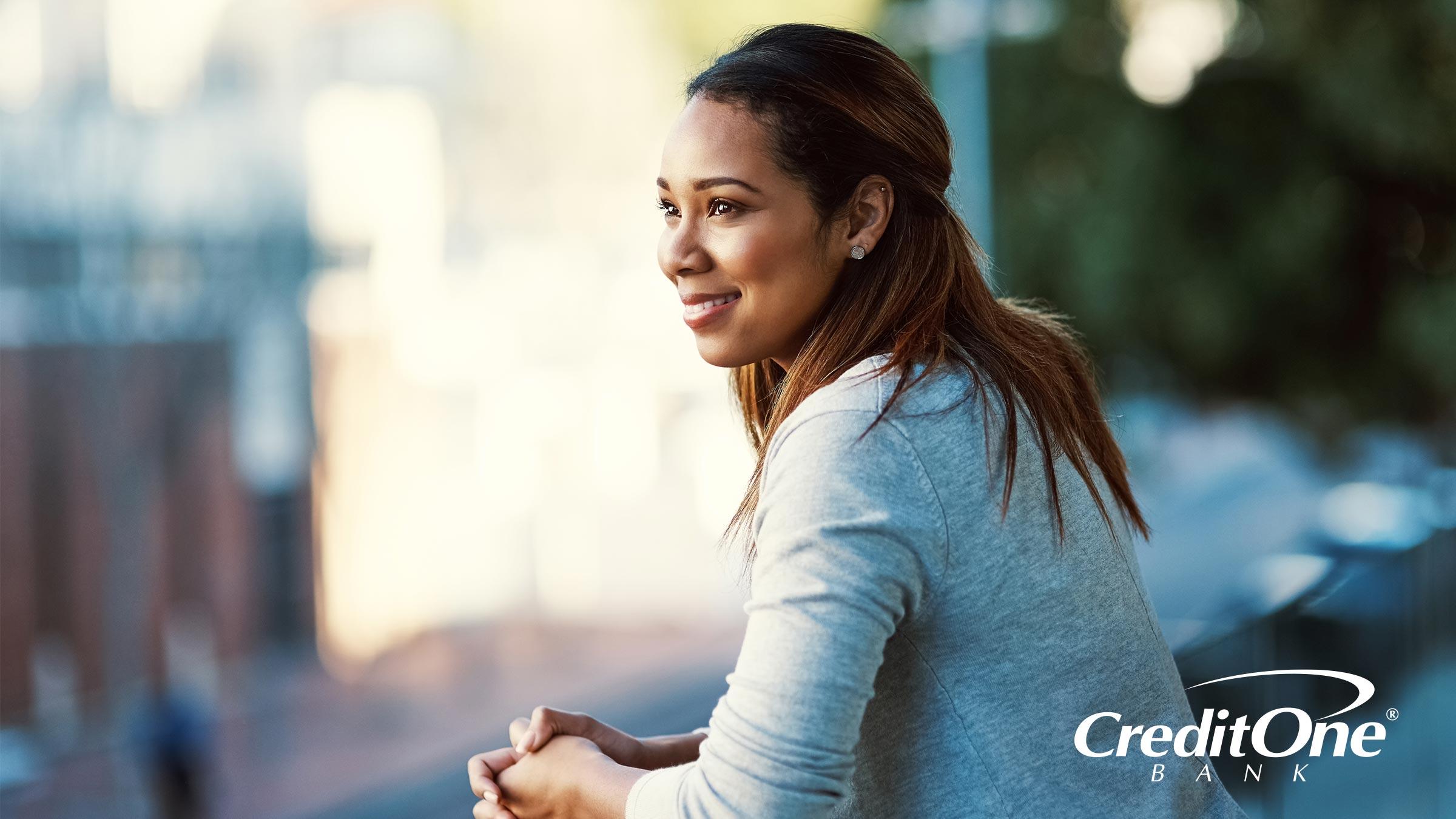 A woman smiles as she gazes off into the distance from a balcony above street level. She looks relieved and accomplished, perhaps because she finally set up her personalized financial alerts.